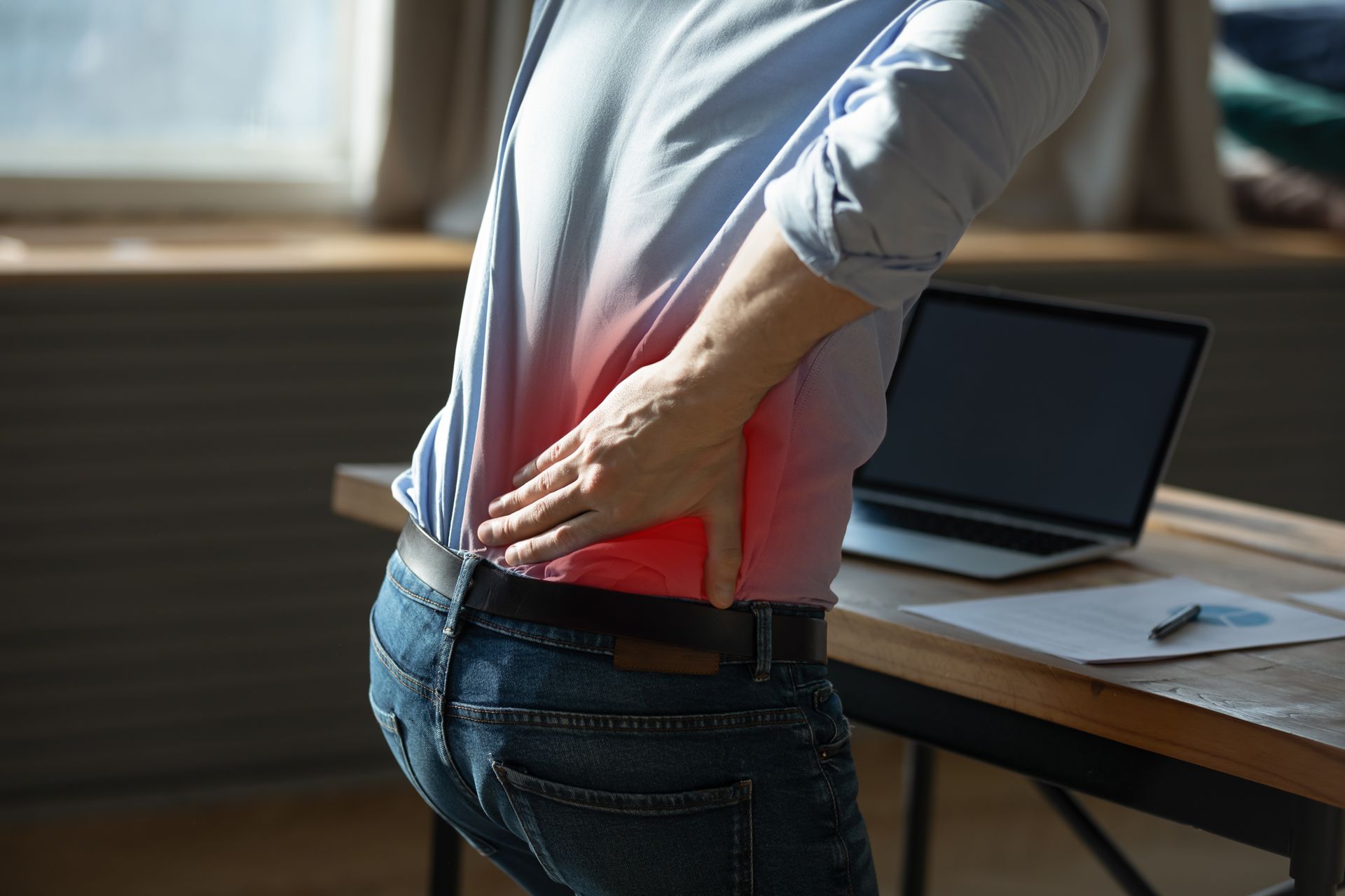 Person with hand on lower back, experiencing back pain near desk and laptop. 