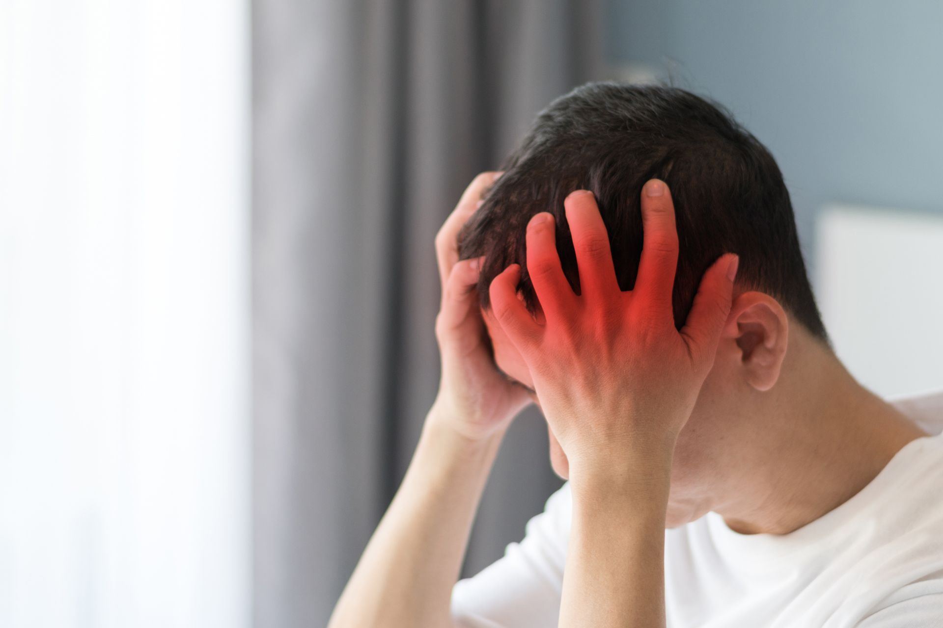 Man holding head, red glow highlights hands, indicating pain. 
