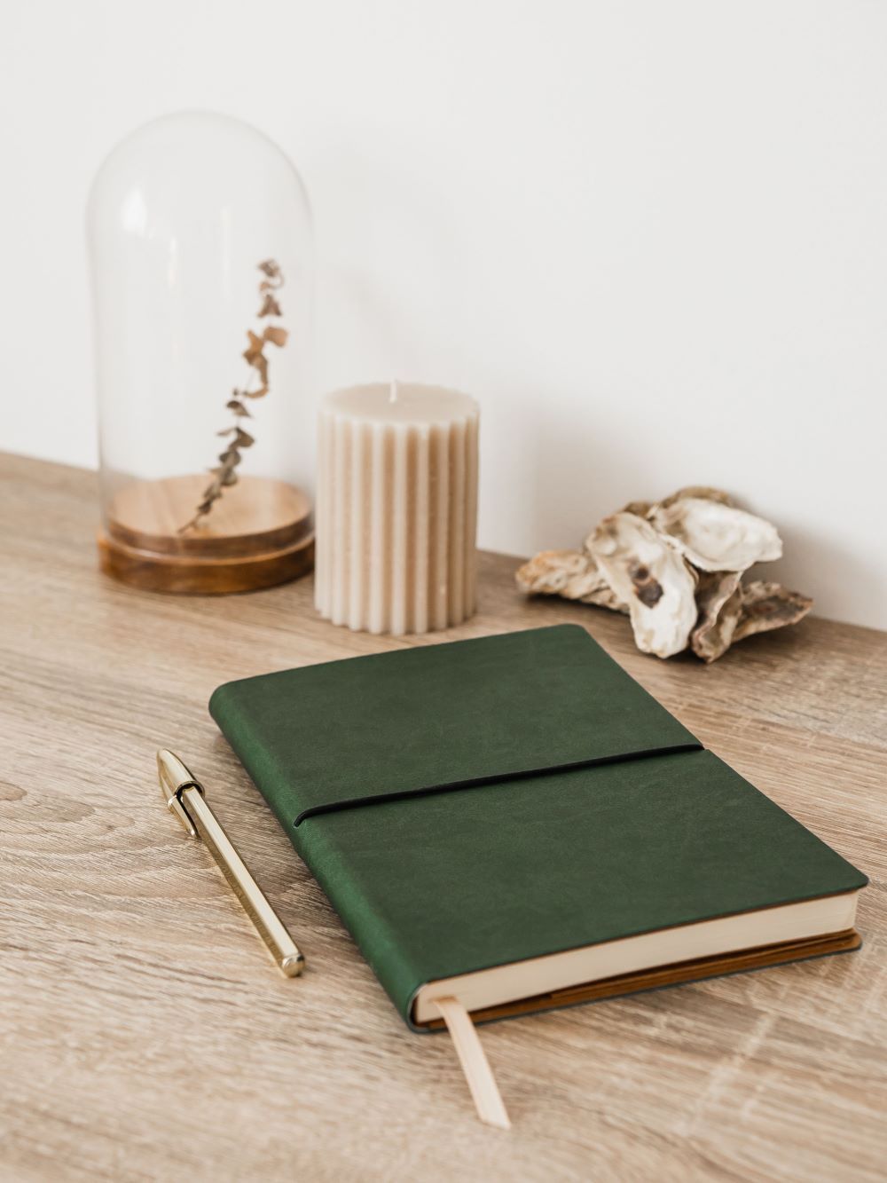 Green notebook and pen on a wooden surface with a candle, dried plant, and seashell.