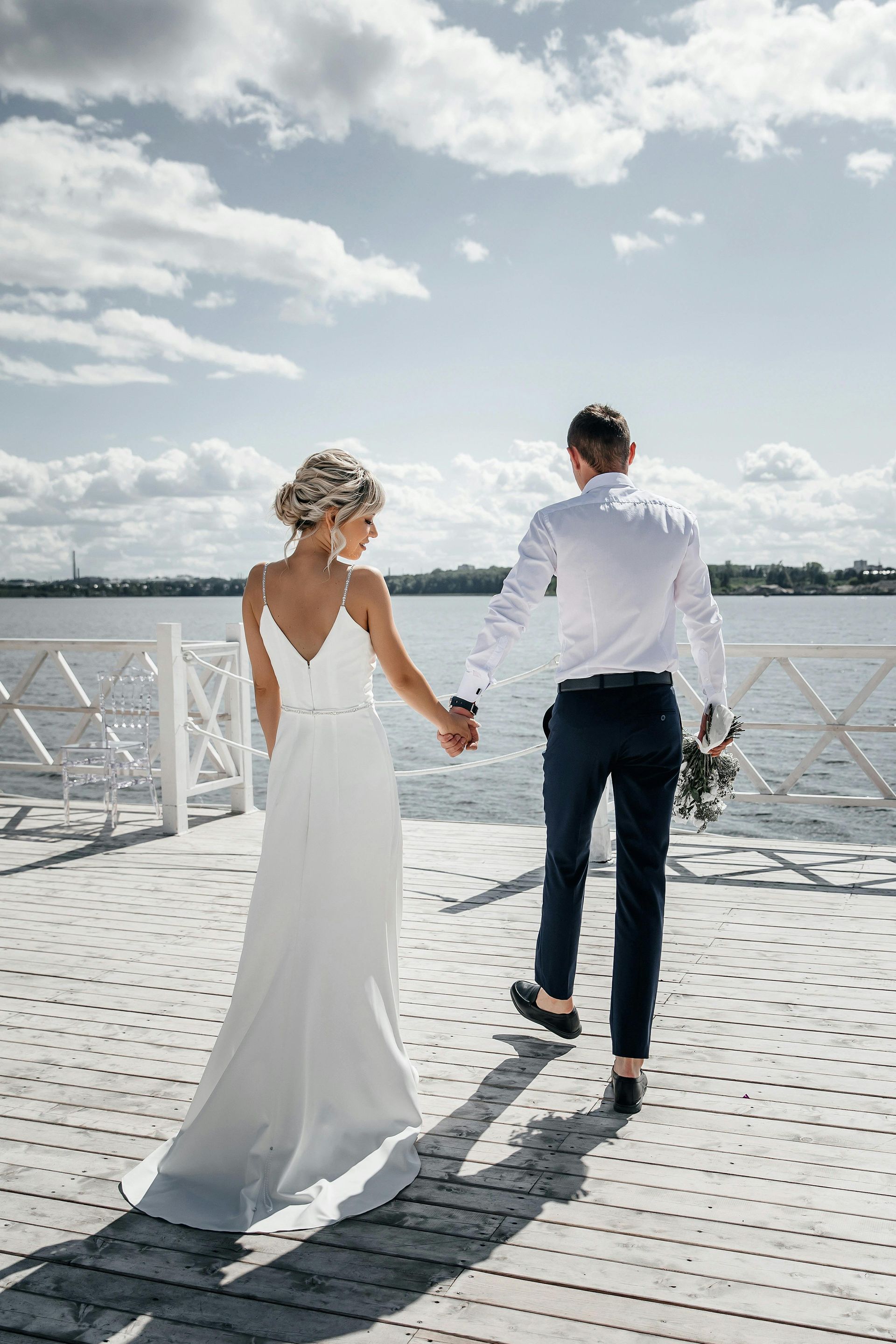 A bride and groom are walking on a pier holding hands.