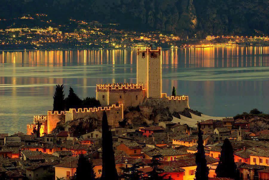 Castello medievale illuminato in riva a un lago al crepuscolo, con le calde luci della città e i riflessi delle montagne sull'acqua.