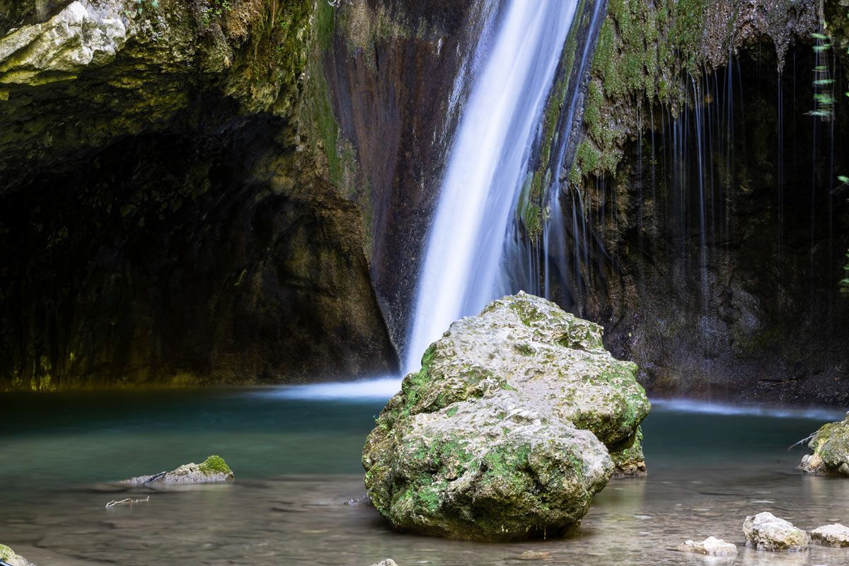 Cascata che si getta in una pozza rocciosa con un masso ricoperto di muschio in primo piano.