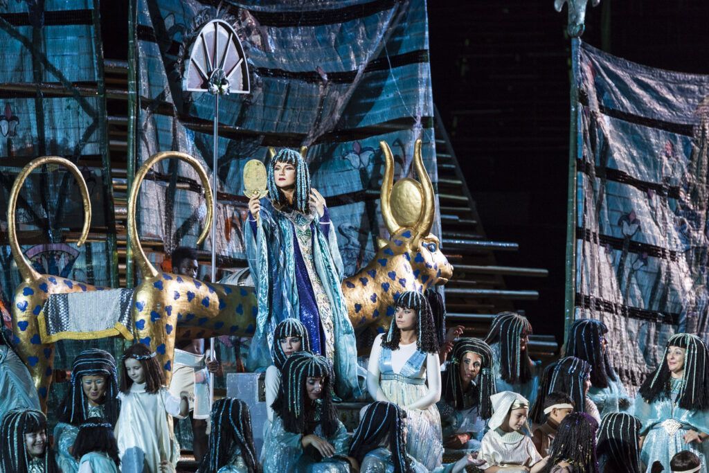 Artisti in costume su un palco illuminato di blu con grandi oggetti di scena dorati e uno sfondo simile a un castello