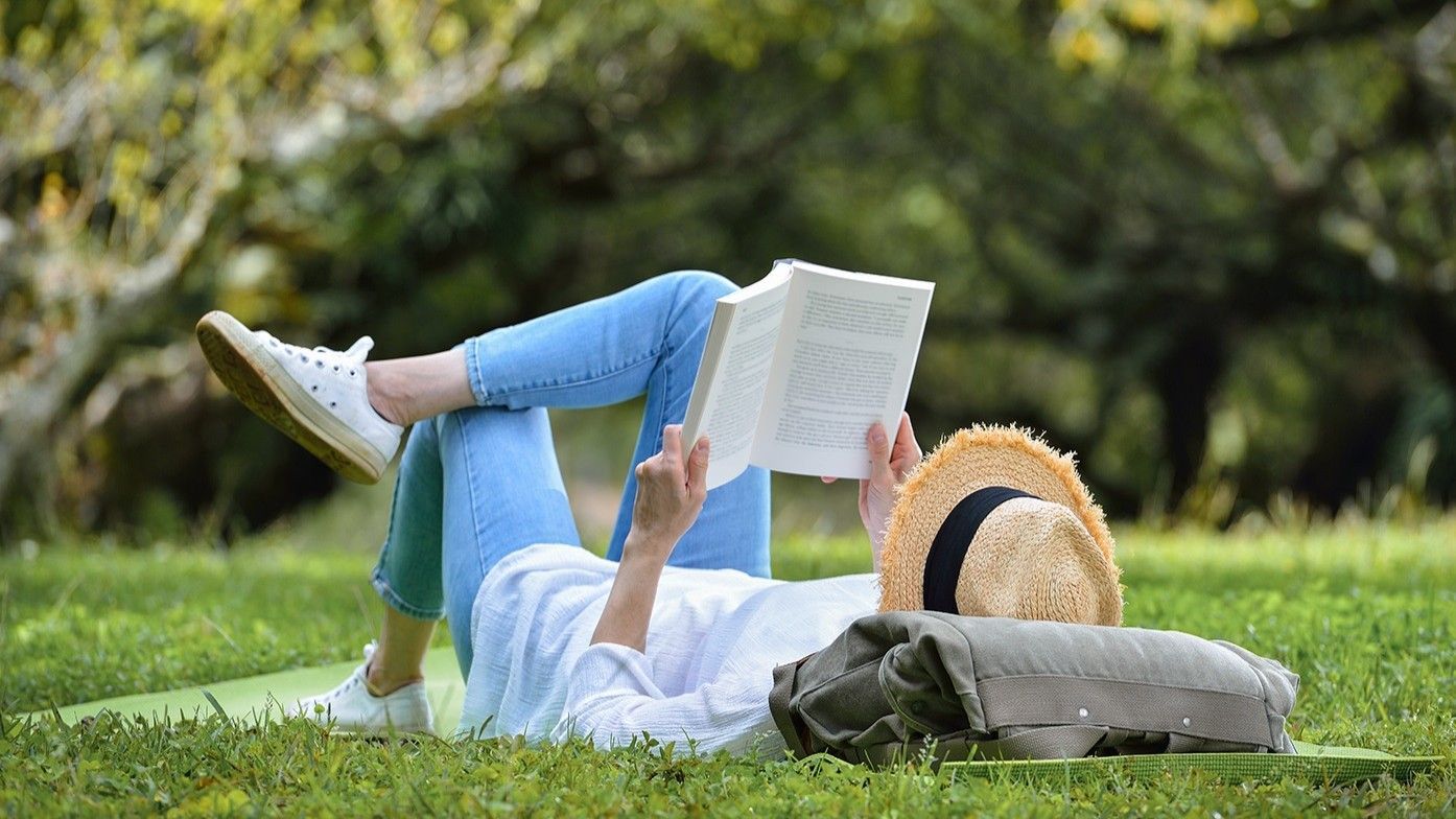 Una persona legge su una coperta in un parco erboso, indossando jeans e una camicia bianca, con un cappello di paglia e dei cuscini nelle vicinanze.