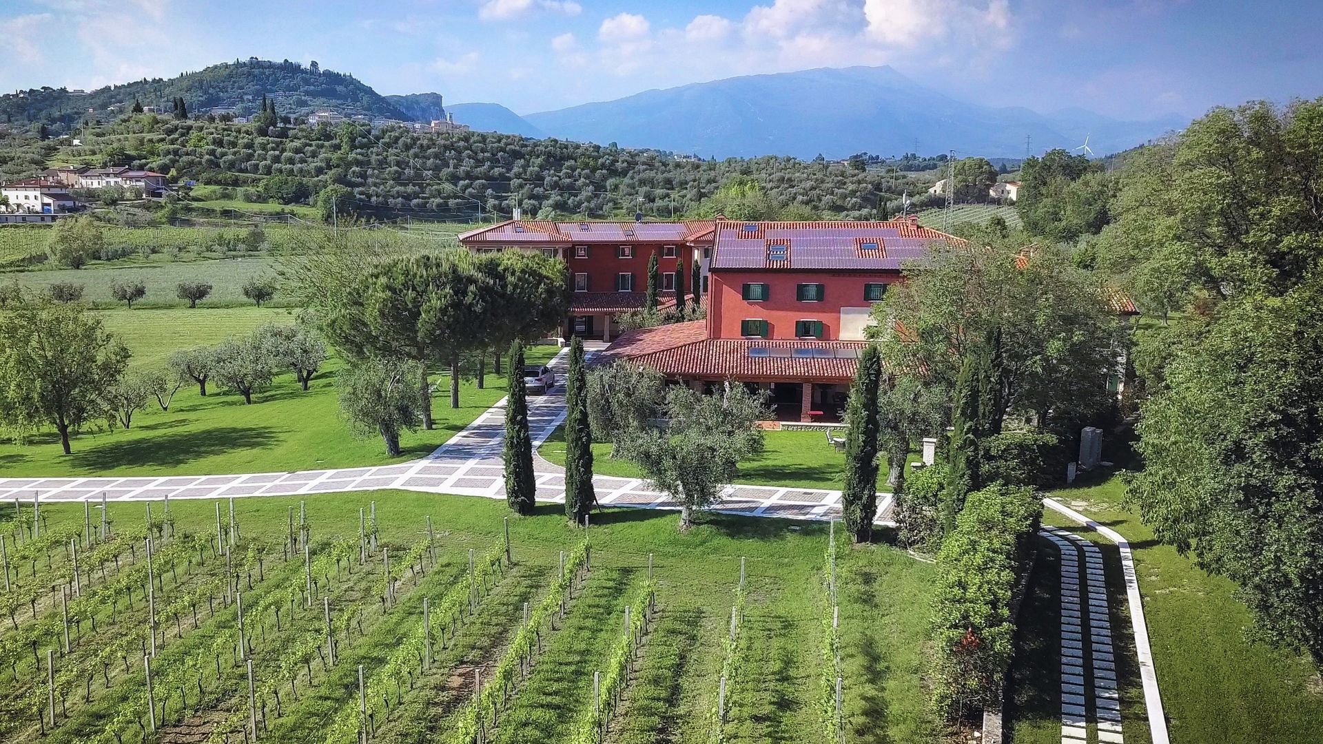 Filari di vigneti che conducono a una casa colonica rossa in una verde vallata con colline e montagne sullo sfondo