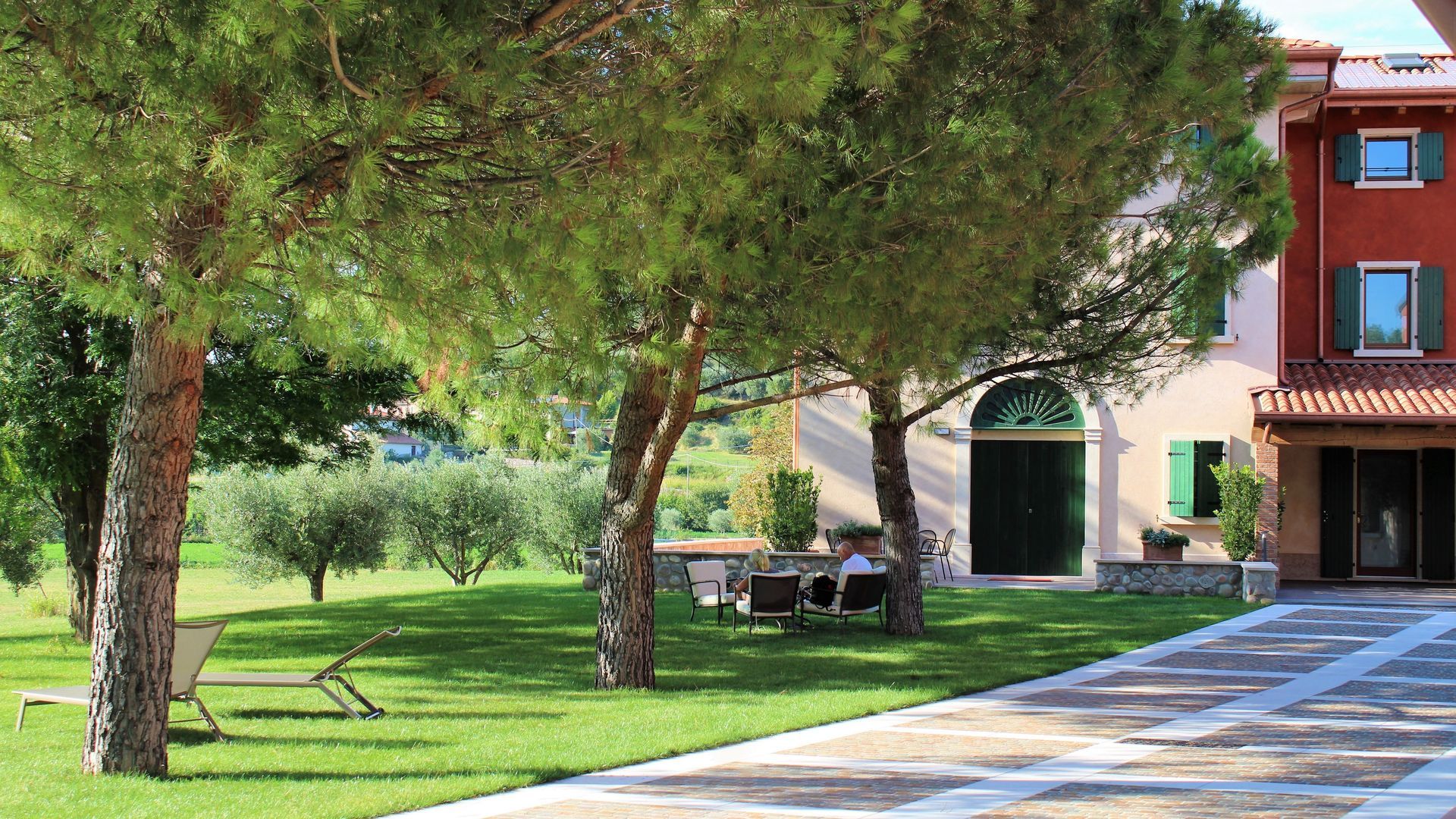 Prato alberato accanto a un edificio moderno con panchine e un vialetto pavimentato.