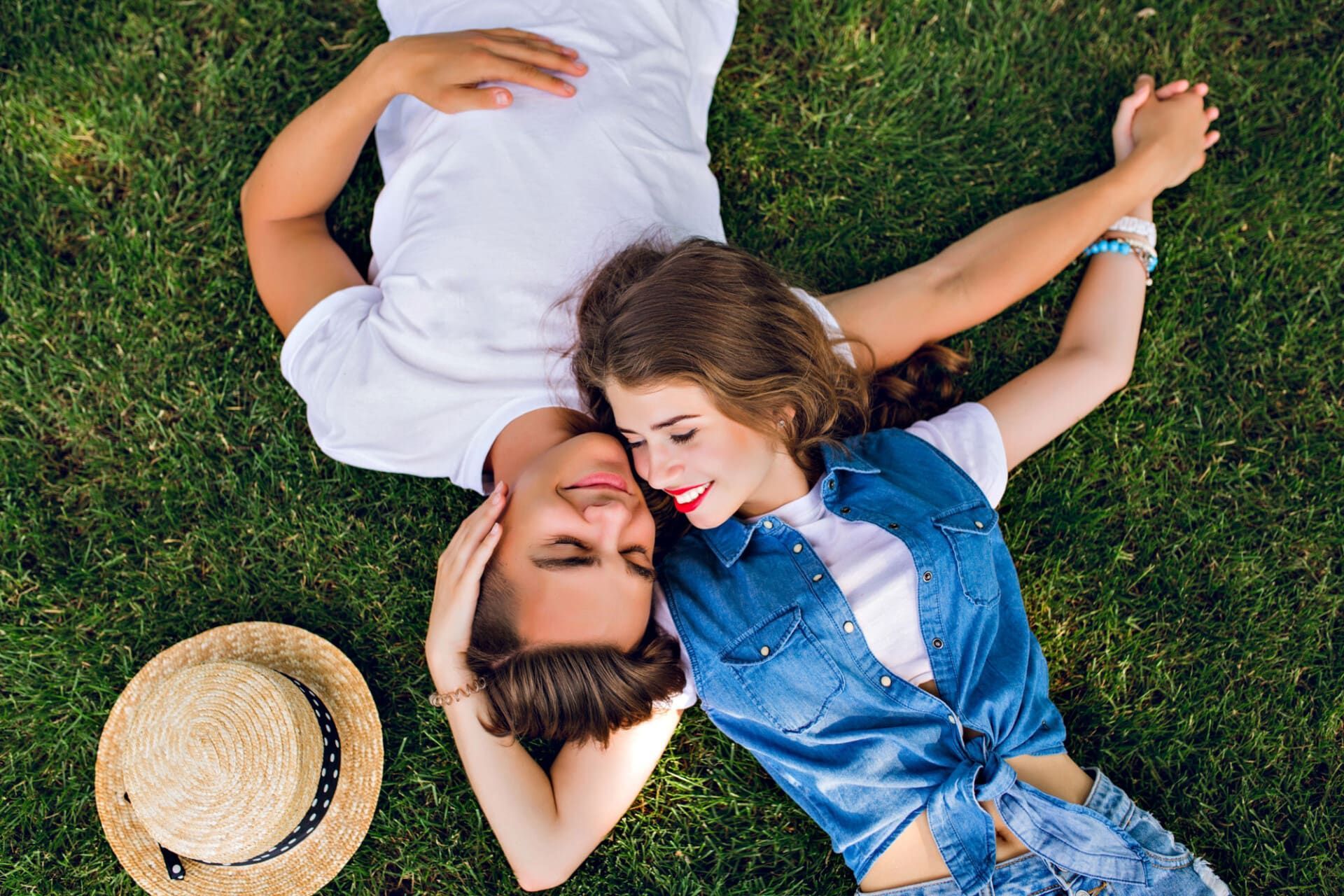 Due persone sdraiate sull'erba con le teste vicine, sorridenti, accanto a un cappello di paglia