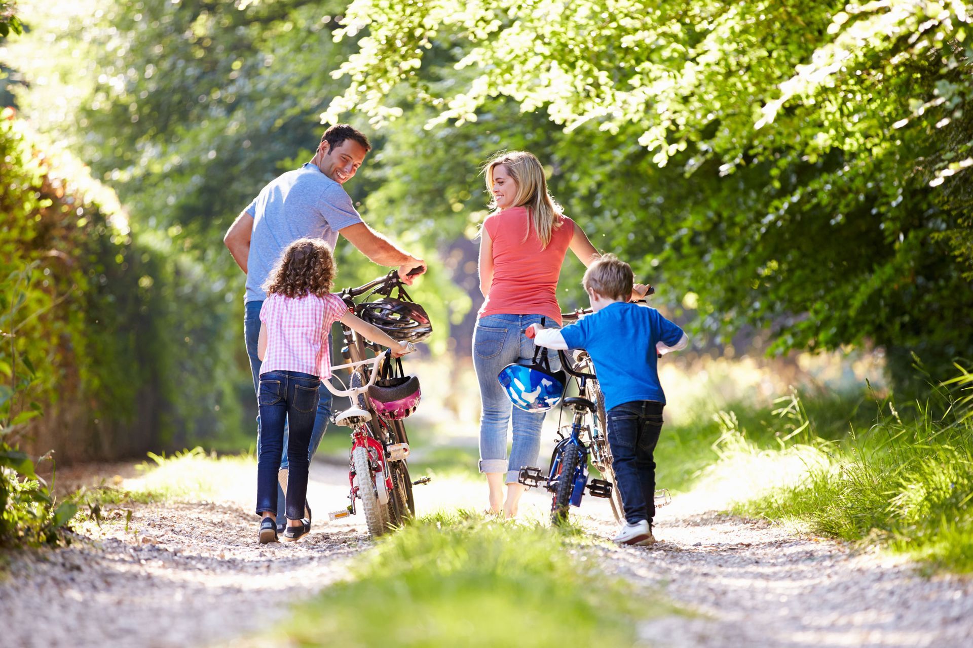 Una famiglia con due bambini e biciclette su un sentiero di ghiaia soleggiato in un parco alberato.