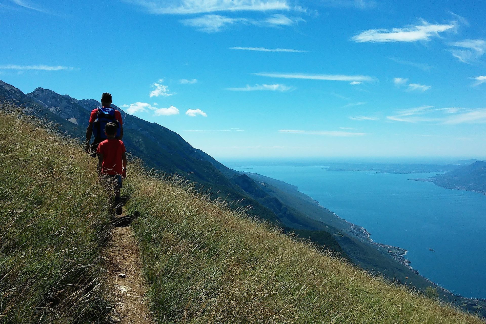 Escursionista con zaino rosso che cammina su un sentiero di montagna erboso, costeggiando un oceano blu e colline in lontananza.