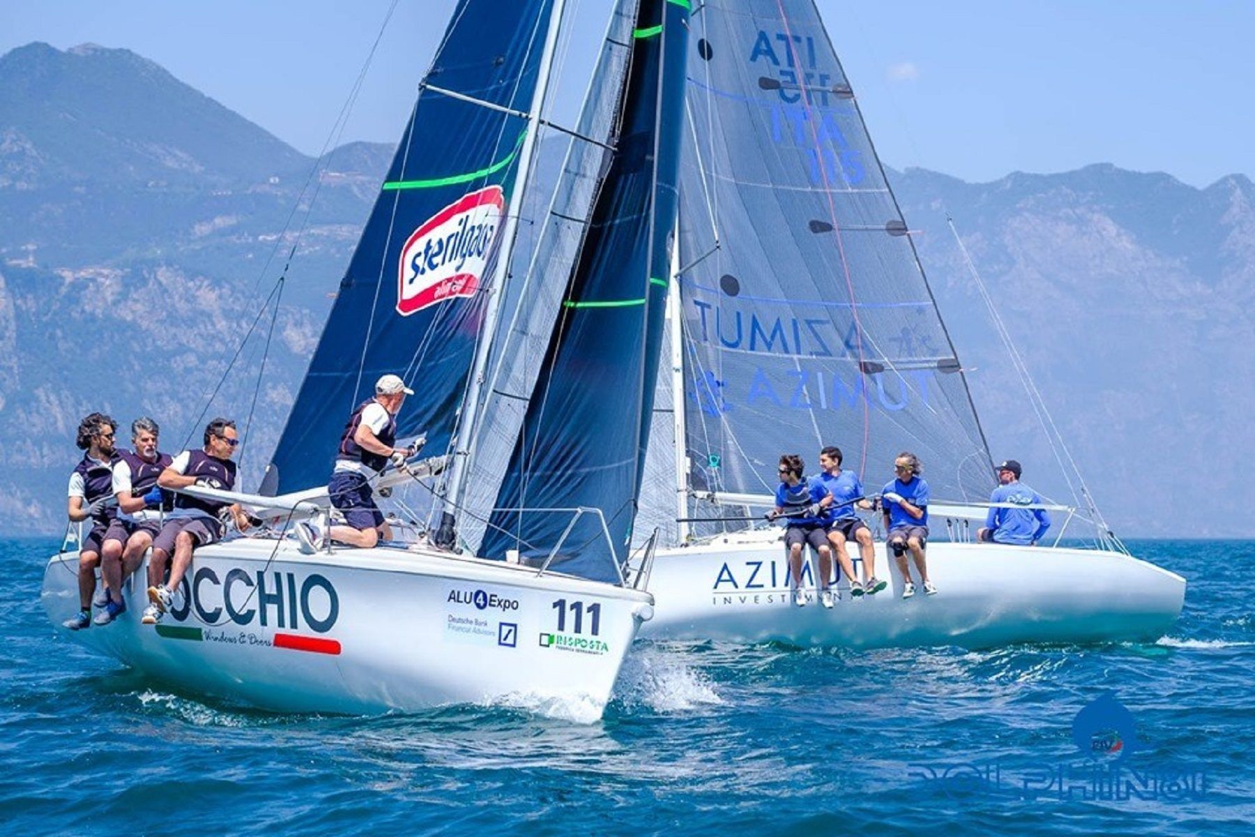 Due barche a vela che regatano su acque blu con l'equipaggio a bordo e le montagne sullo sfondo.
