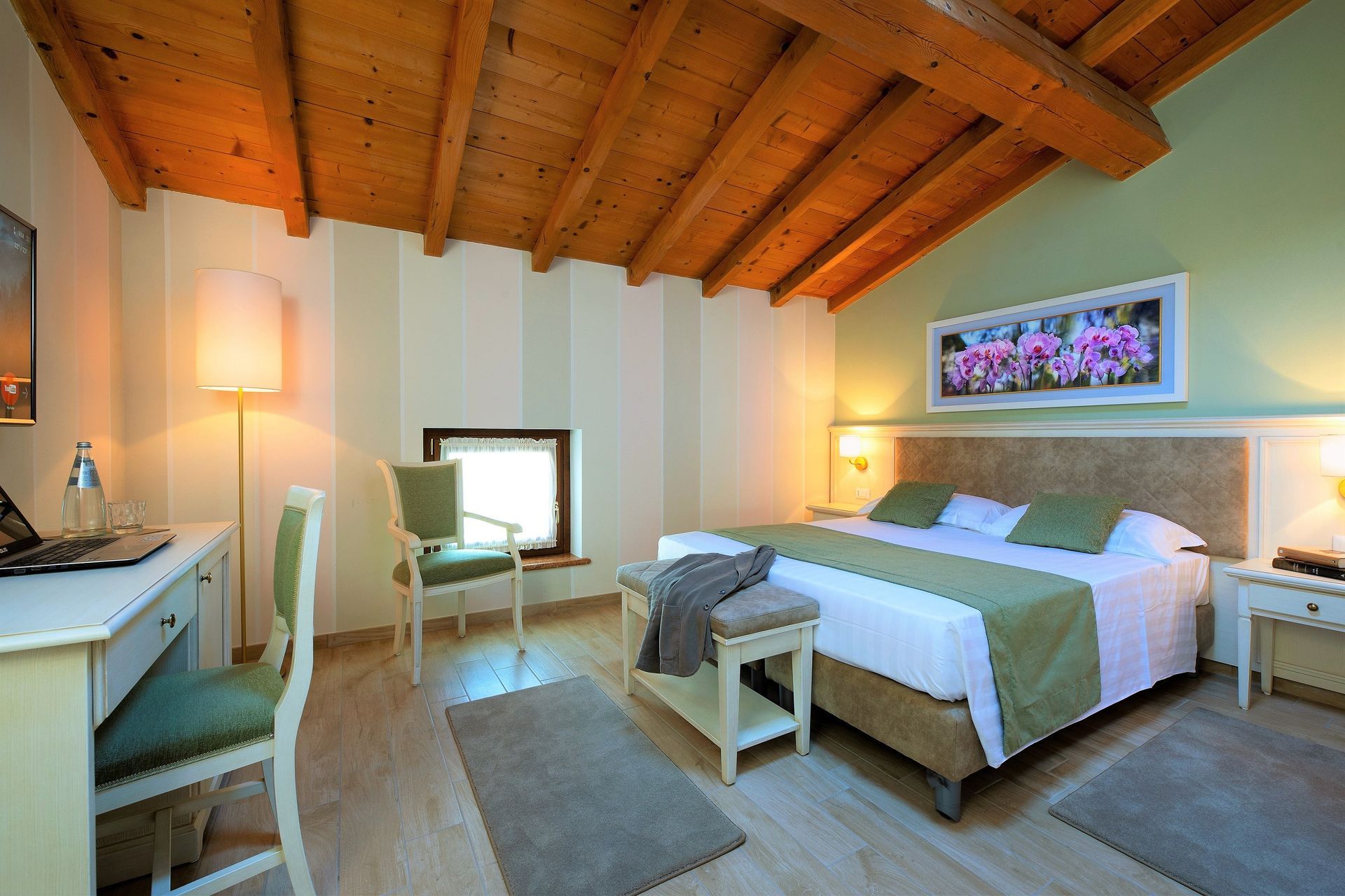 Accogliente camera da letto con soffitto in legno, letto bianco, scrivania, lampada e quadro floreale sopra la testiera.