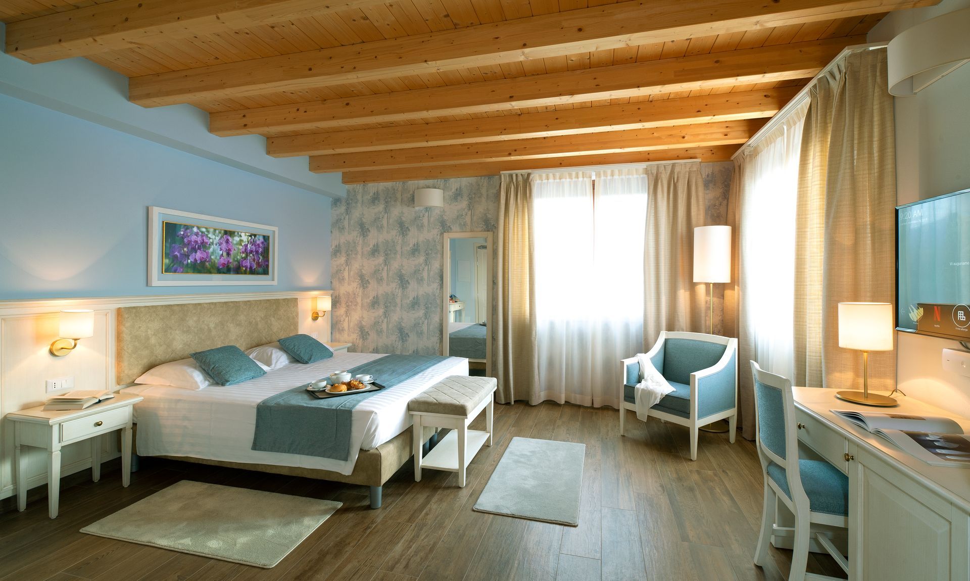 Accogliente camera da letto con travi in ​​legno a vista, letto, sedie, scrivania e morbida luce naturale proveniente dalle tende.