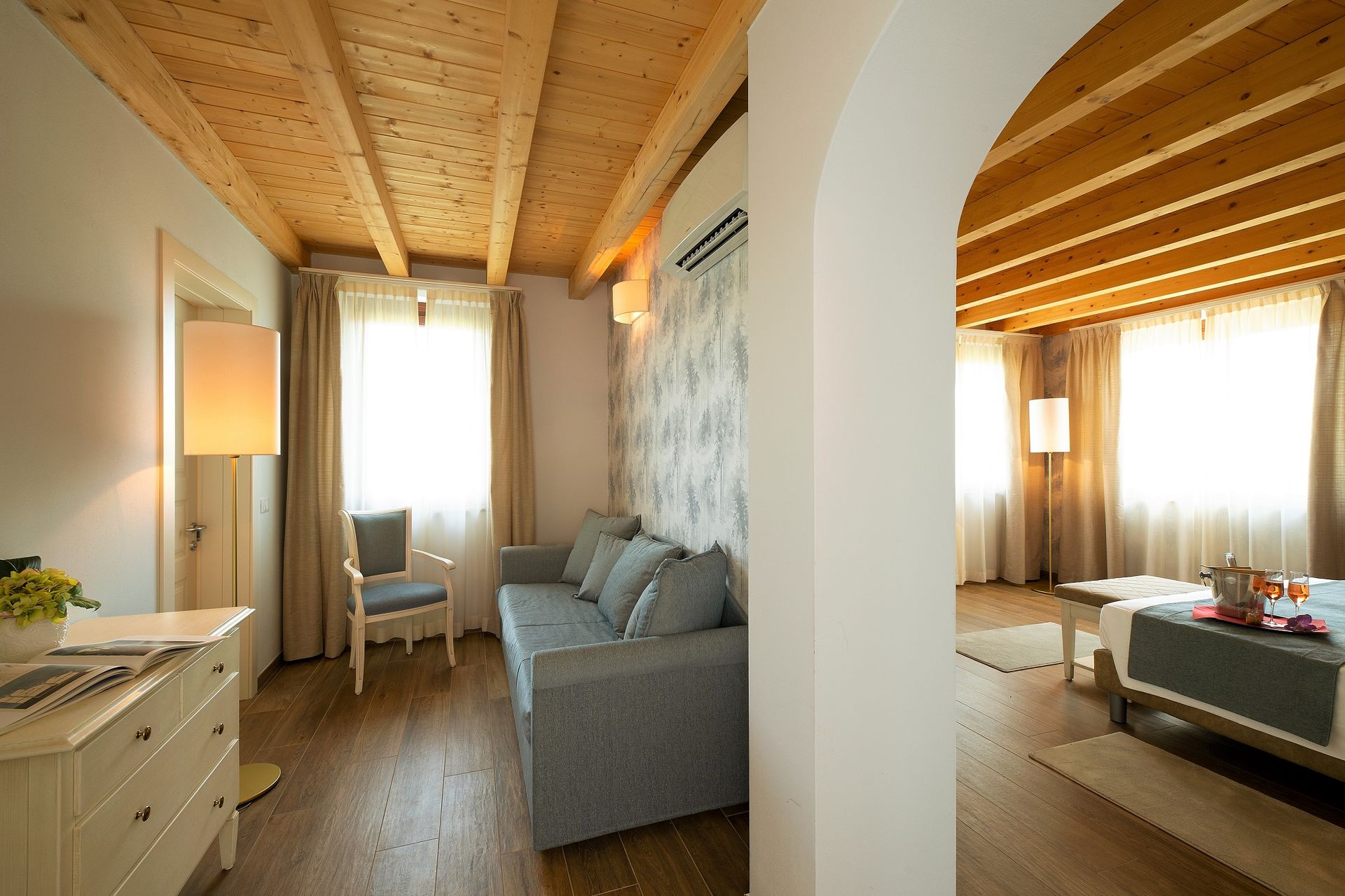 Camera da letto luminosa e accogliente con travi a vista sul soffitto, divano, scrivania e luce naturale che filtra attraverso le tende.