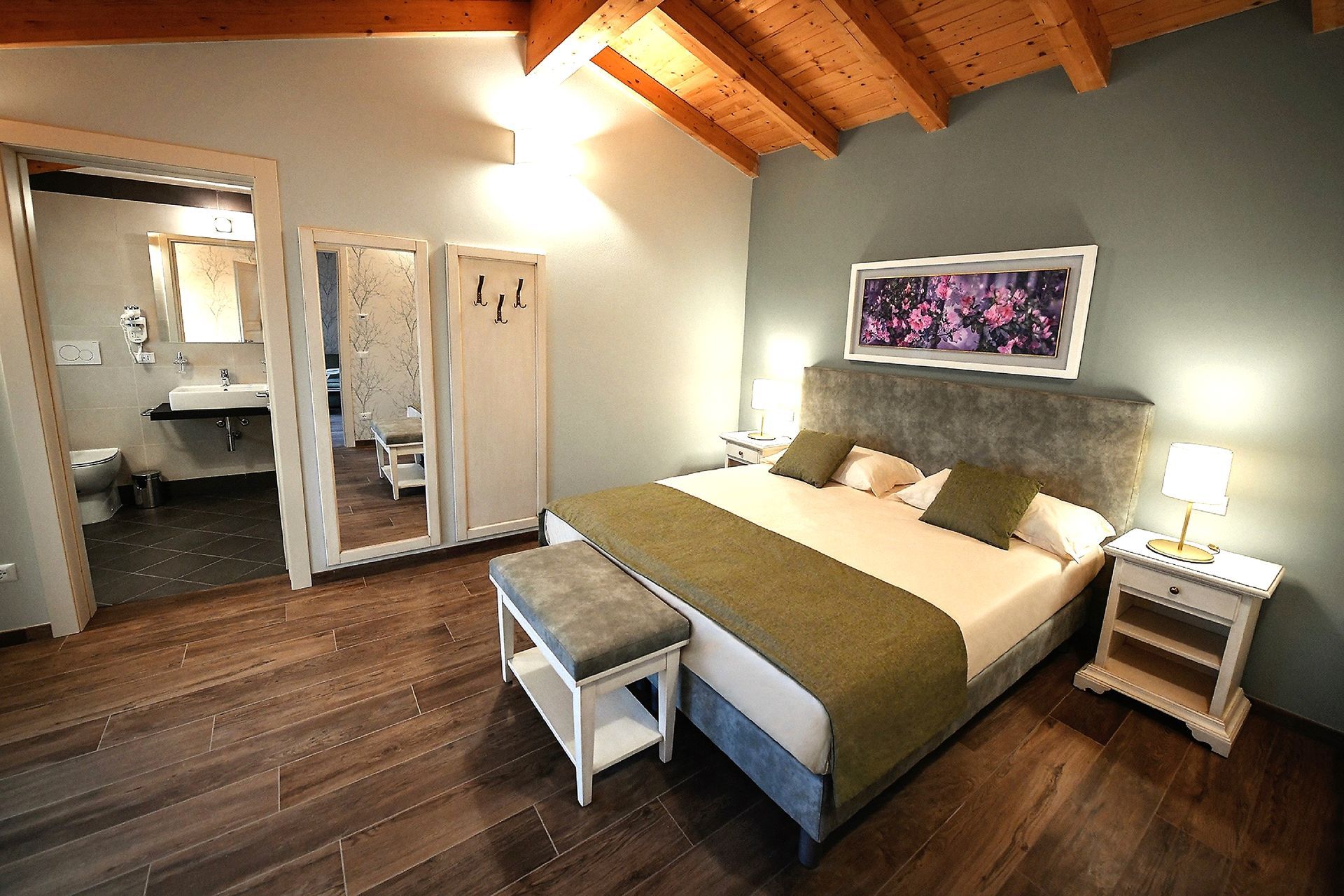 Camera da letto moderna con soffitto in legno, letto king size, lampade da comodino, panca e porte del bagno aperte.