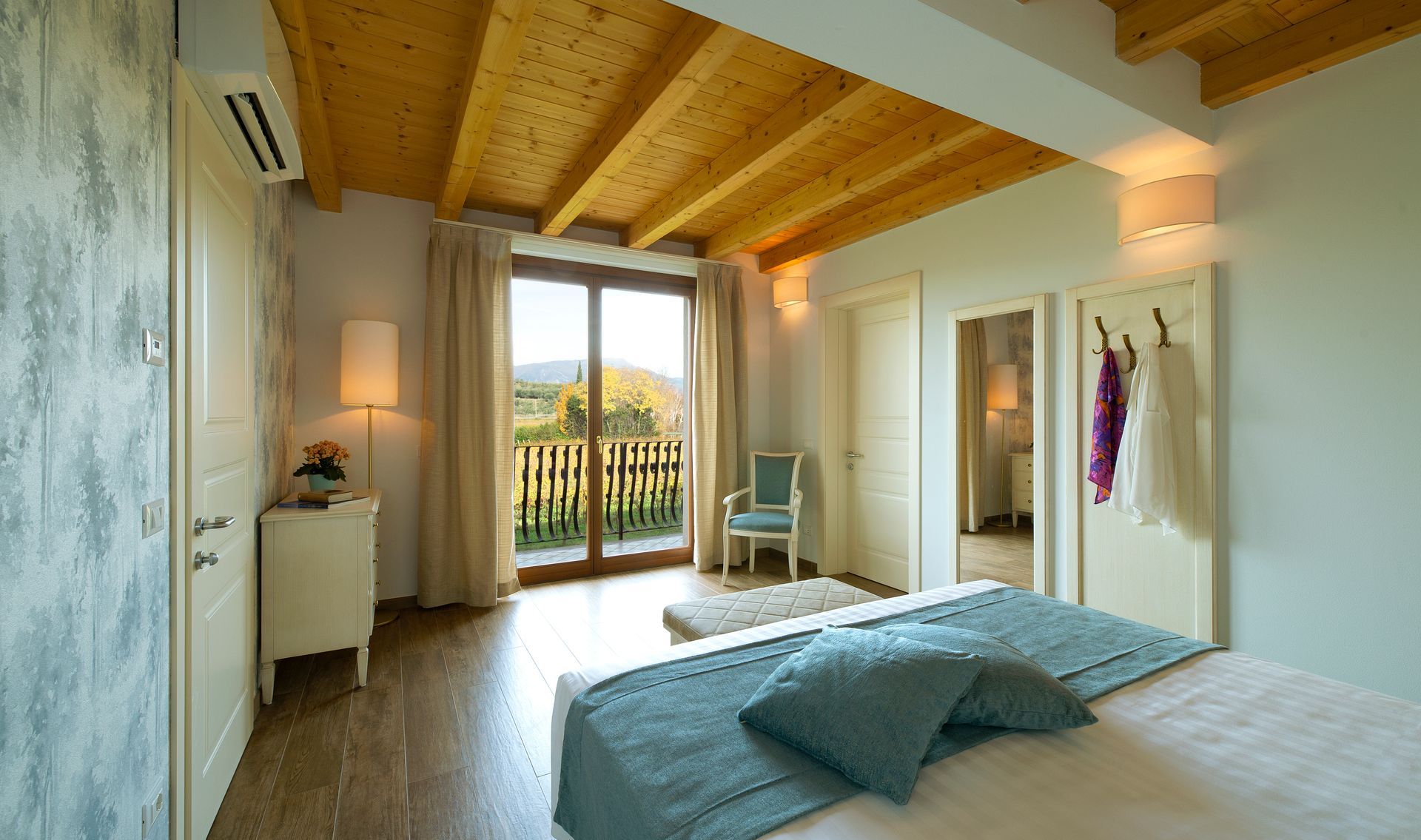 Luminosa camera da letto con soffitto con travi a vista, due letti, ampie porte finestre che danno sul balcone e calda luce naturale.