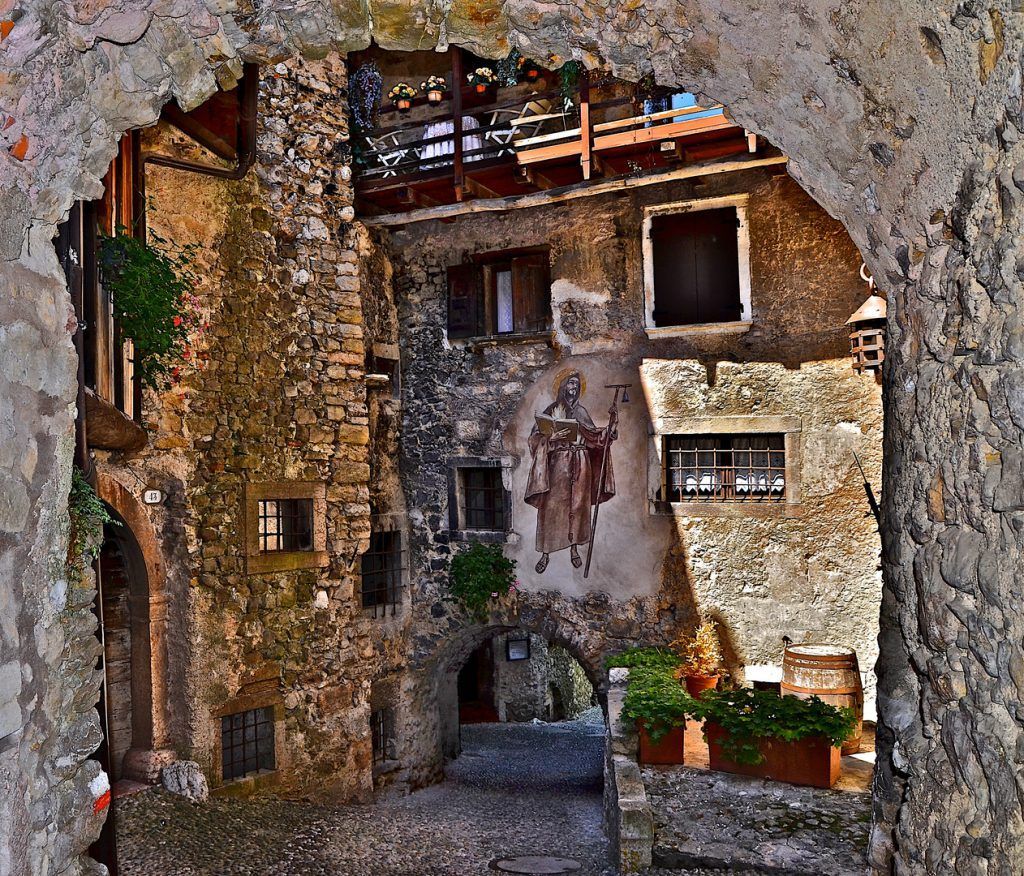 Cortile in pietra incorniciato da un arco, con edifici rustici e vasi di fiori immersi nella calda luce del sole.