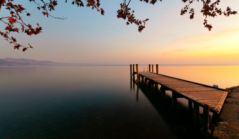 Un molo di legno si protende in acque calme all'alba, incorniciato dalle foglie autunnali.