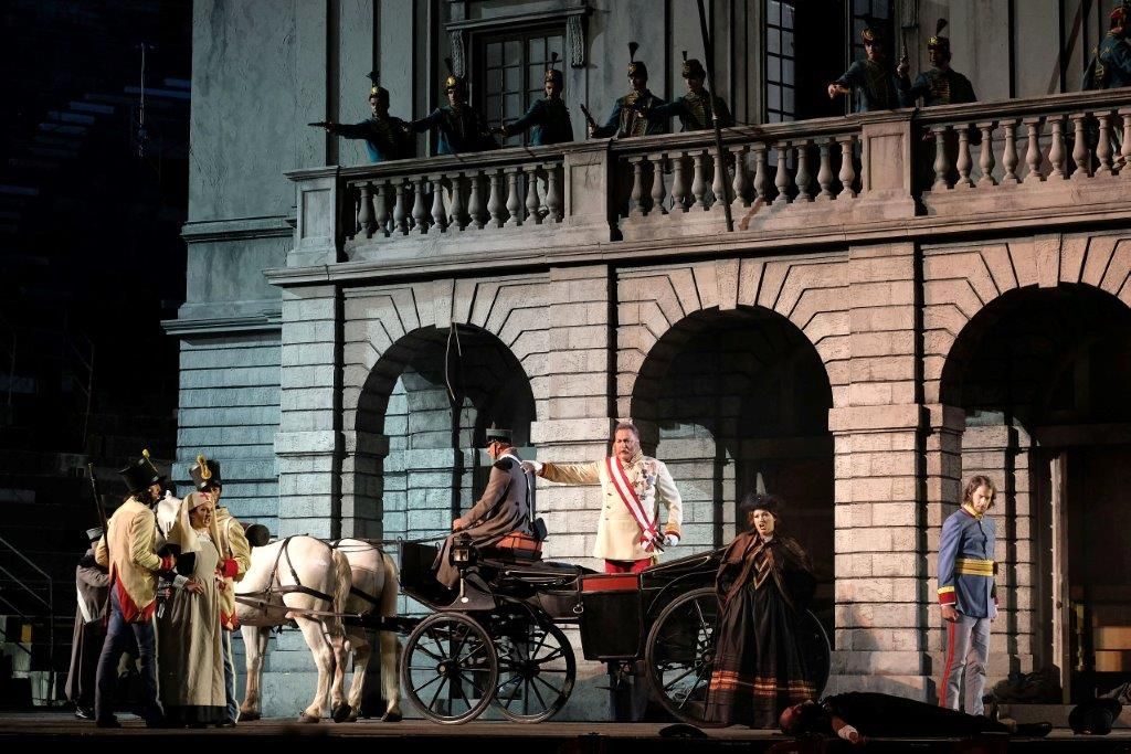 Una carrozza trainata da cavalli si trova davanti a un edificio in pietra ad arco, con persone radunate su una facciata che ricorda un palcoscenico.