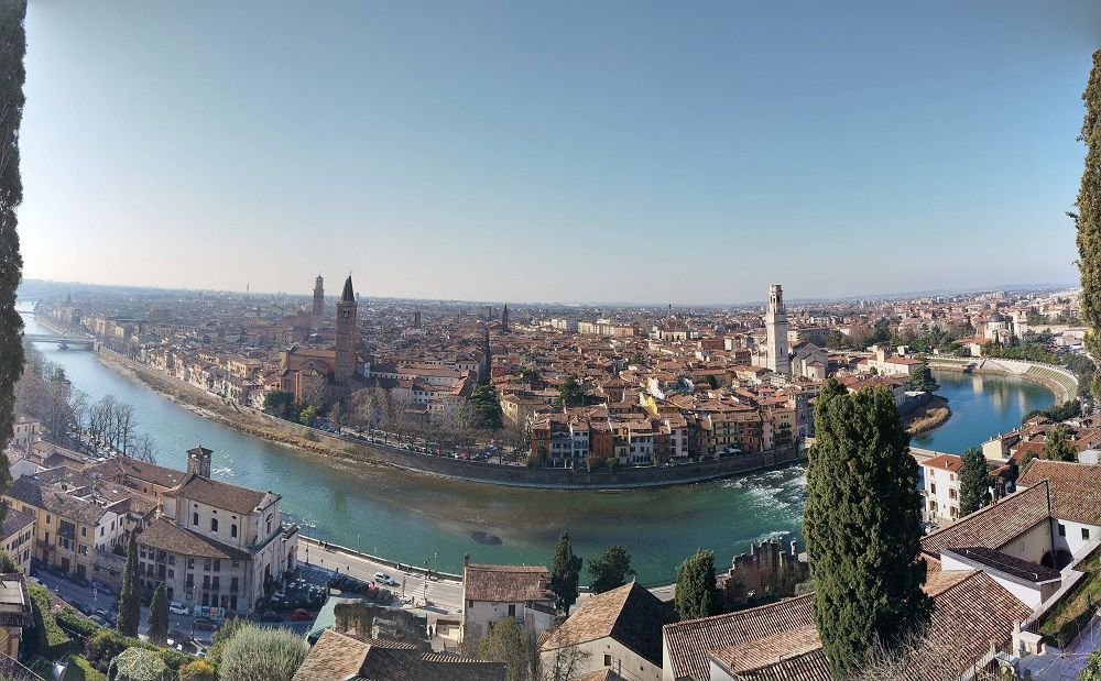 Veduta panoramica di una città storica con un fiume serpeggiante, ponti ed edifici dai tetti rossi sotto un cielo azzurro limpido.