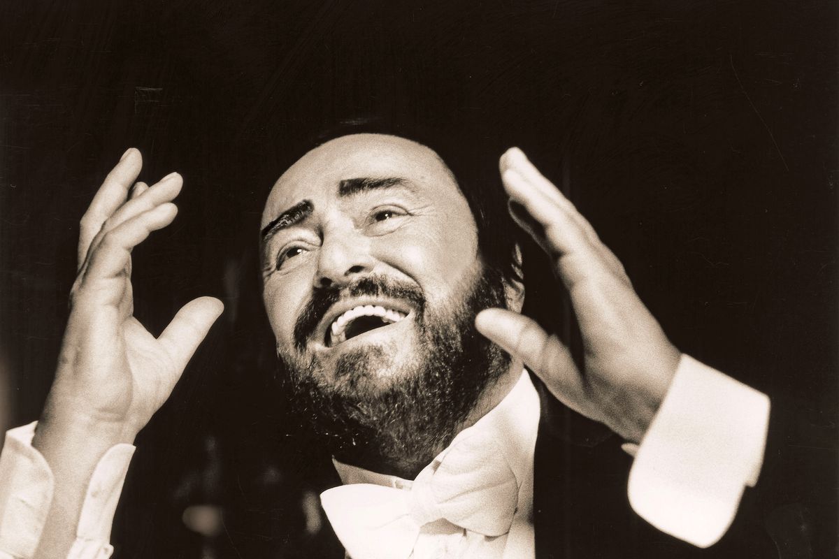 Uomo in smoking con la barba, sorridente e gesticolante con le mani alzate su sfondo scuro
