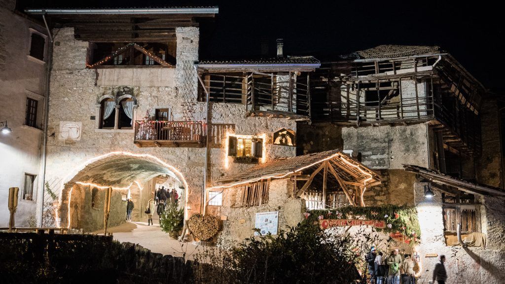 Veduta notturna di un rustico edificio in pietra con luci soffuse, ingresso ad arco e balconi in un villaggio collinare.
