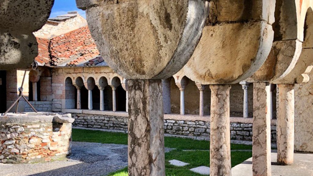 Chiostro in pietra con colonne ad arco, tetto di tegole e cortile soleggiato