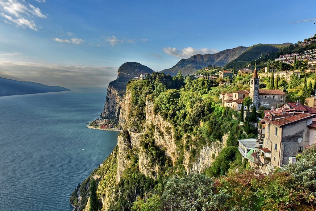 Villaggio a picco sul mare, affacciato su un lago azzurro, con montagne illuminate dal sole e case a schiera sotto un cielo limpido.