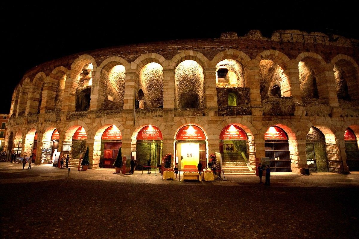 Anfiteatro di pietra illuminato di notte, con aperture ad arco e persone radunate al di sotto.