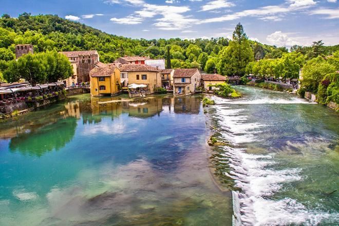 Fiume che scorre tra case rustiche e vegetazione sotto un cielo azzurro brillante