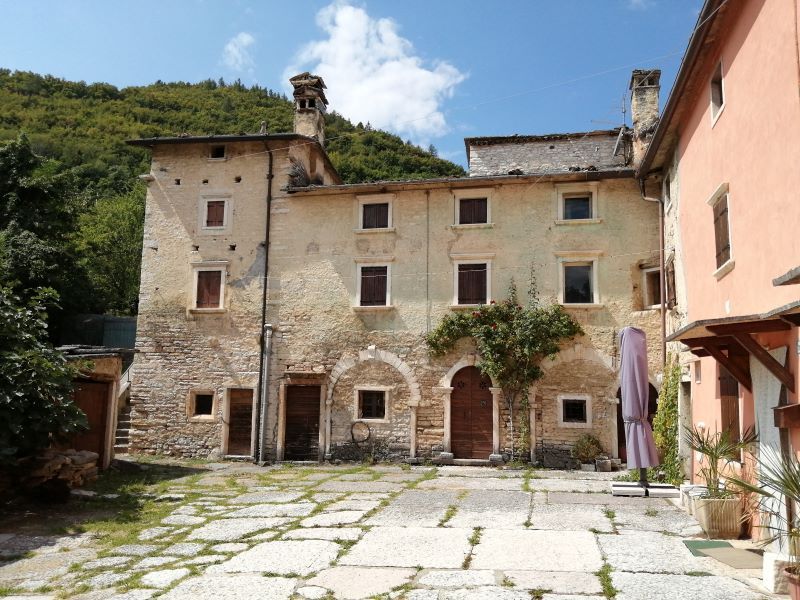 Cortile in pietra con vecchi edifici beige, portali ad arco e una collina sotto un cielo azzurro.