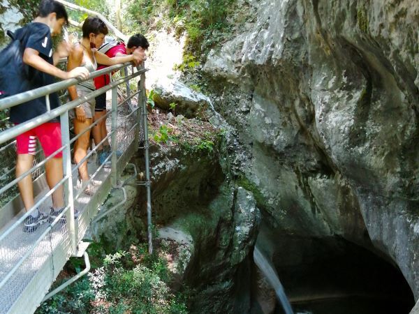 Alcune persone si sporgono da una ringhiera, scrutando l'apertura di una grotta rocciosa accanto a un sentiero a strapiombo.
