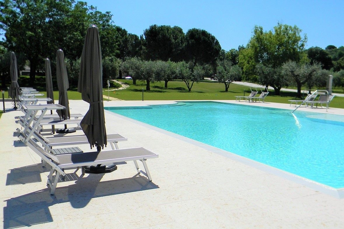 Piscina all'aperto con lettini, ombrelloni chiusi e alberi verdi sotto un cielo azzurro limpido.