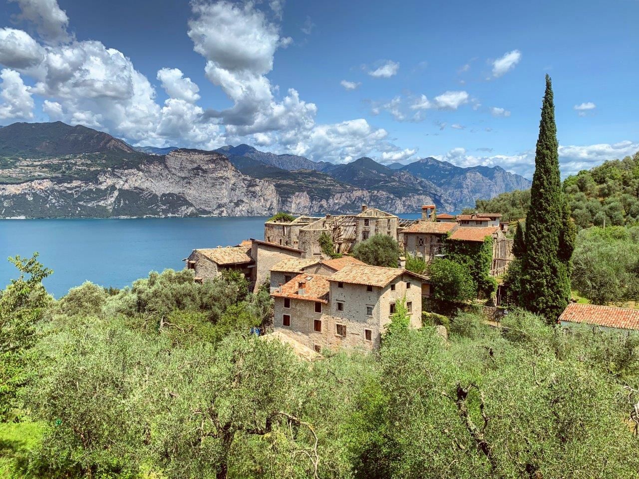 Villaggio sul lago con case dai tetti di terracotta, ulivi e montagne sotto un cielo azzurro nuvoloso.