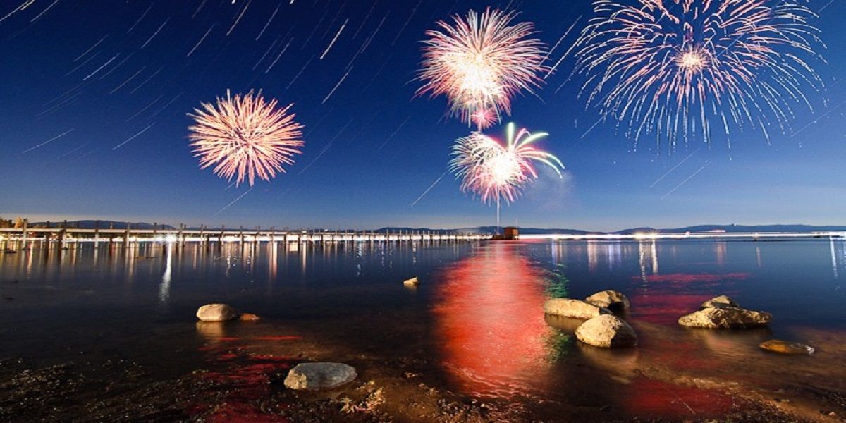 Fuochi d'artificio su una città lacustre di notte, che si riflettono sull'acqua calma e sugli scogli della riva.