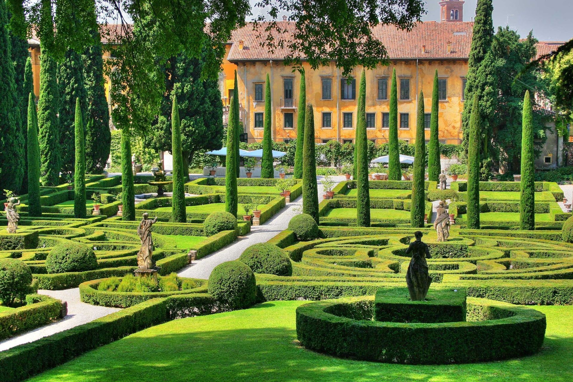 Giardino formale con siepi ben curate, alberi ad alto fusto e una villa sullo sfondo.