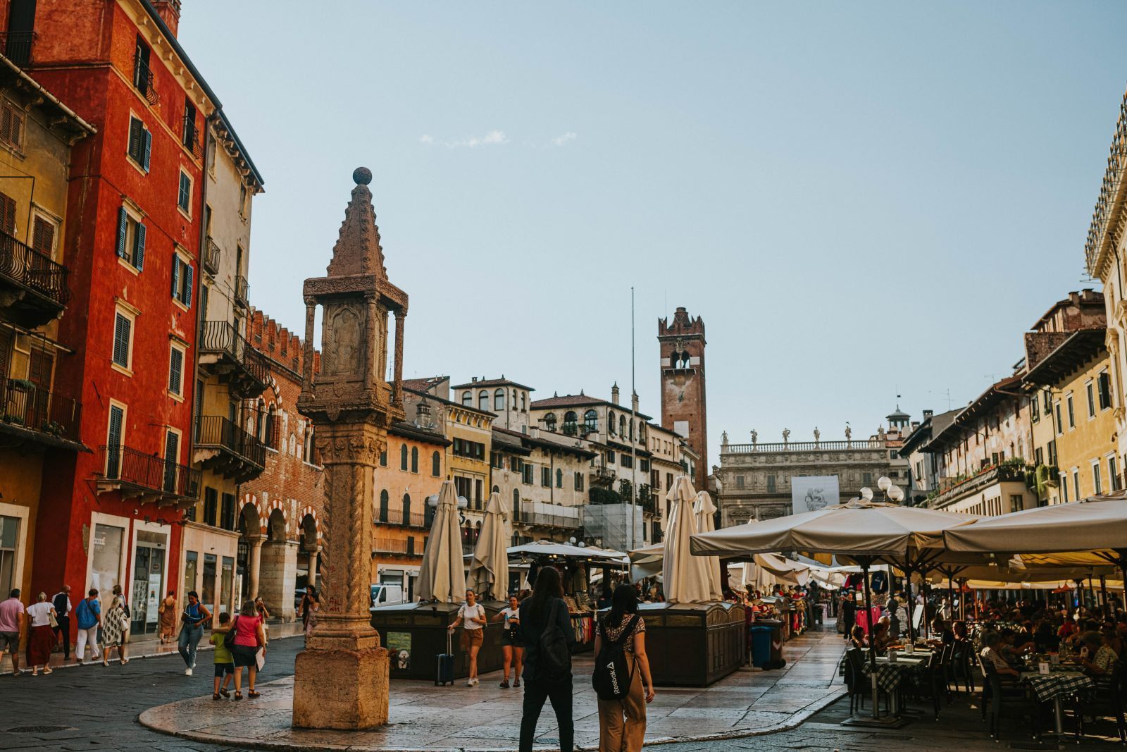 Piazza storica europea con edifici in pietra, un monumento centrale e persone che camminano su una strada acciottolata e bagnata.
