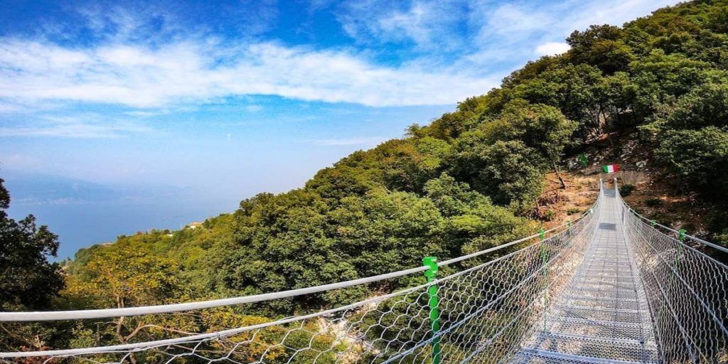 Un ponte sospeso attraversa un pendio boscoso sotto un cielo azzurro, con un escursionista in lontananza che lo percorre.