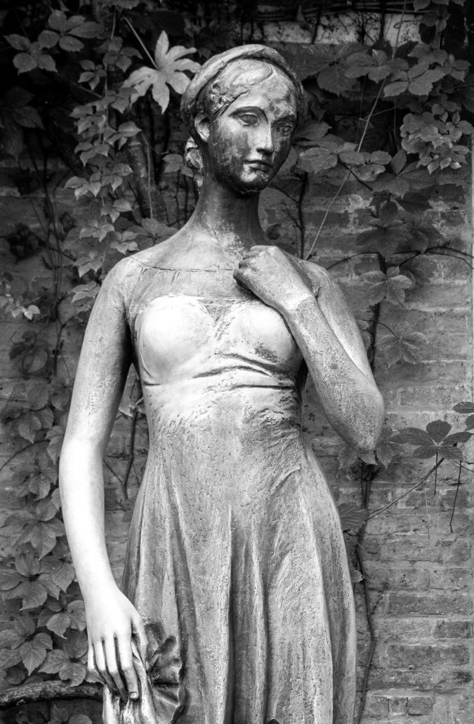 Statua in bianco e nero di una donna drappeggiata in piedi contro un muro di mattoni, con una mano appoggiata sul petto.