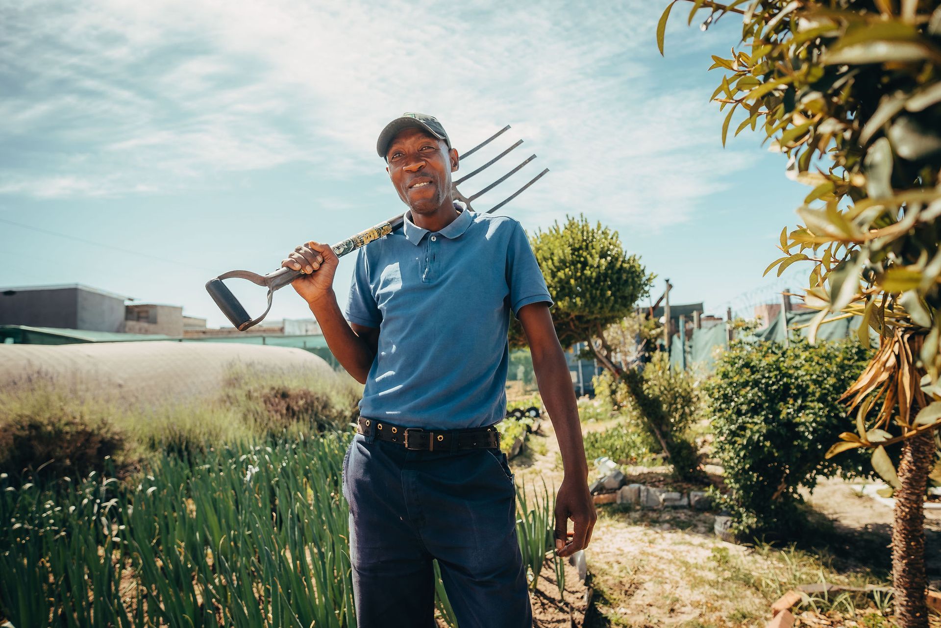 Een man met een hooivork over zijn schouder in een tuin, gekleed in een blauw shirt en een pet. Zonnige dag.