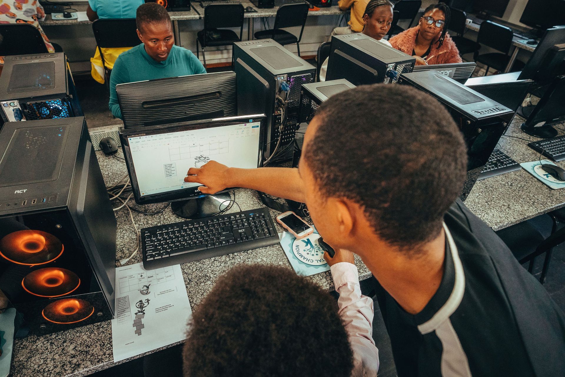 Mensen in een computerlokaal, sommigen kijken naar beeldschermen. Iemand wijst naar een scherm en helpt anderen.