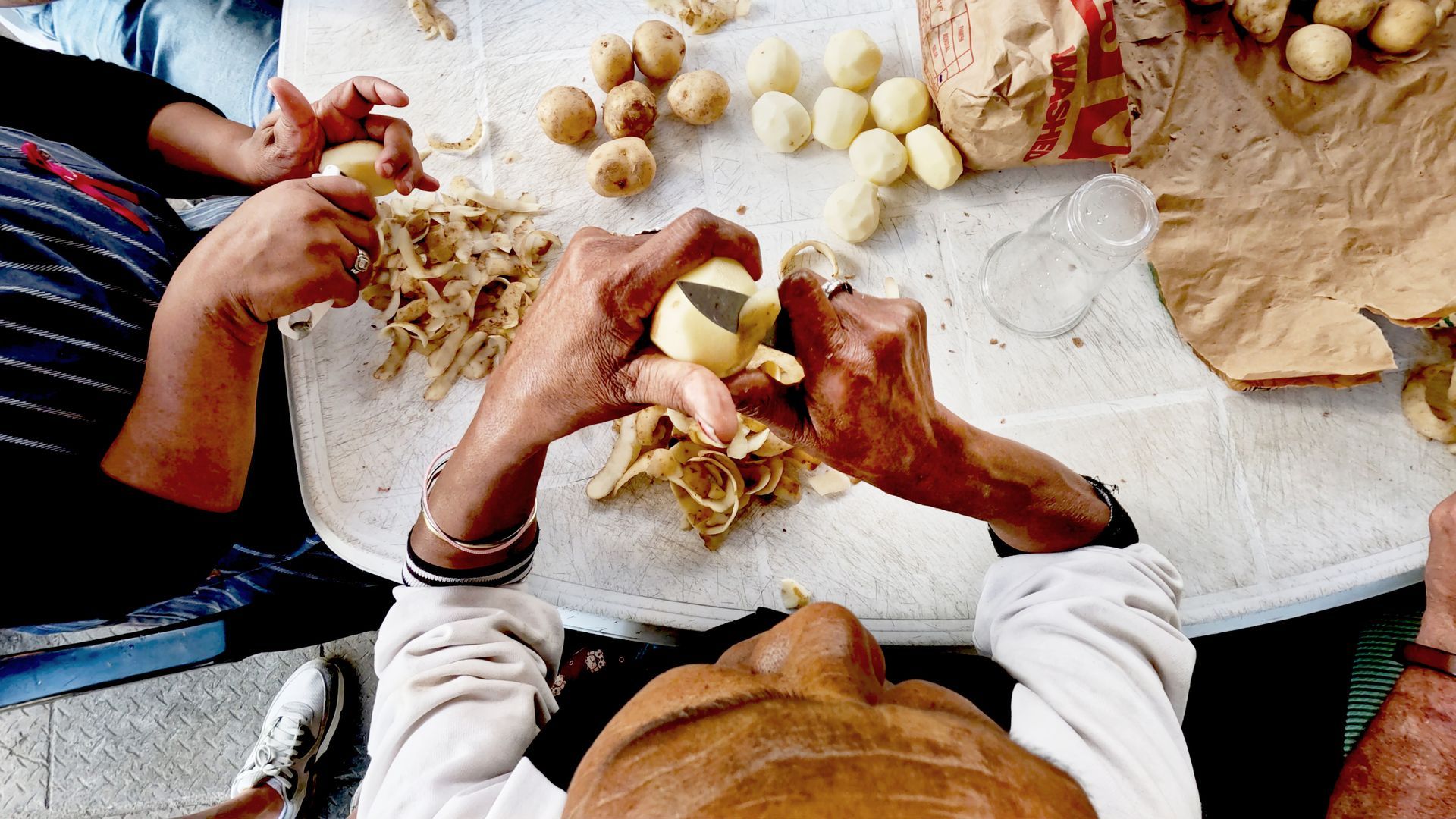 Handen die aardappelen schillen op een witte ondergrond, waarbij de geschilde aardappelen en de schillen zichtbaar zijn.