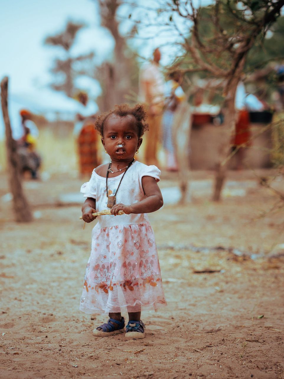 A little girl in a white dress is standing in the dirt.