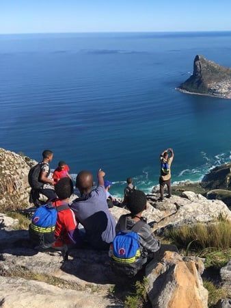 A woman is talking to a group of children sitting on the ground.