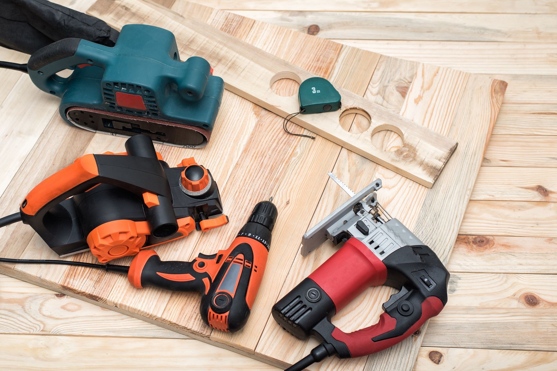 Assorted power tools arranged on a wooden surface beside drilled boards and accessories.