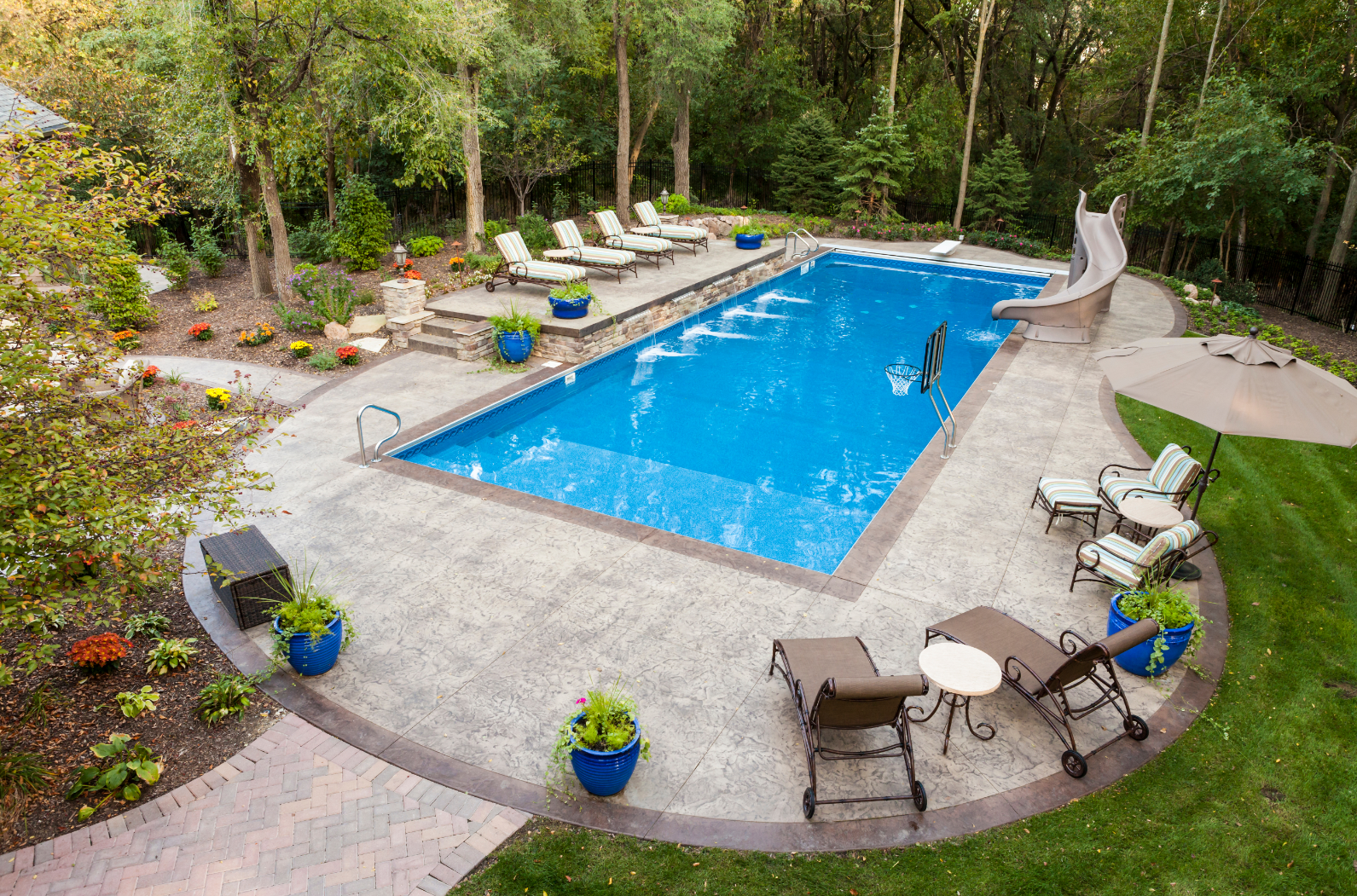 A backyard pool with a concrete deck surrounding it.  There a a number of lounge chairs and a slide.
