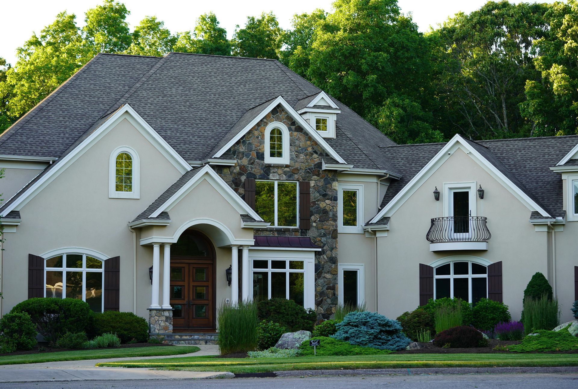 Two-story beige house with stone accents, arched windows, and brown shutters, set amidst lush greenery.
