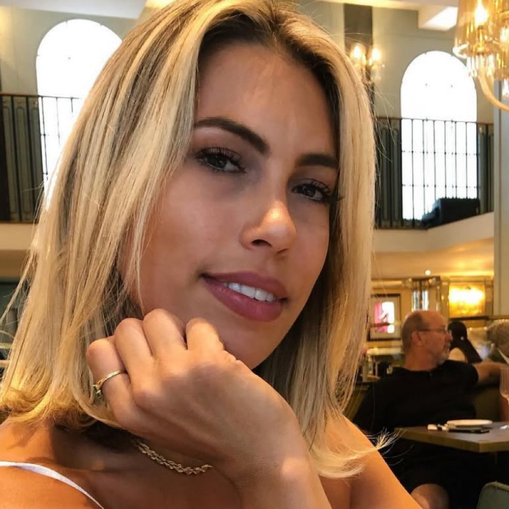 Woman with blonde hair, smiling, resting chin on hand. Restaurant setting with other people.