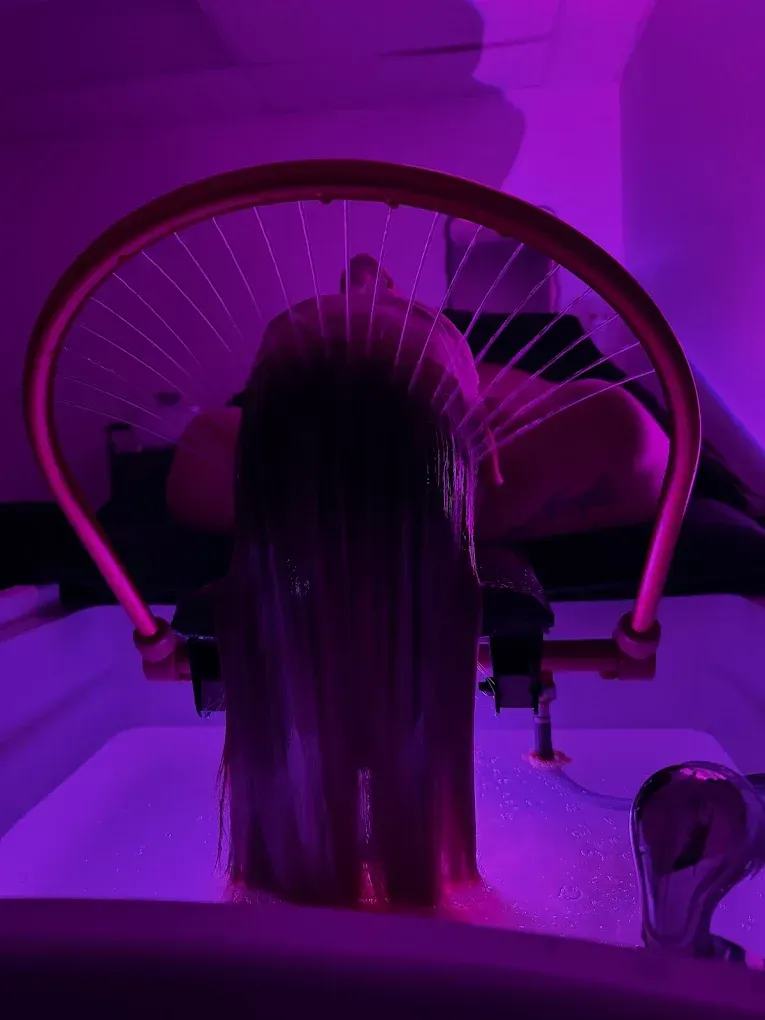 Woman's hair wet under a salon shower head, illuminated by purple light.
