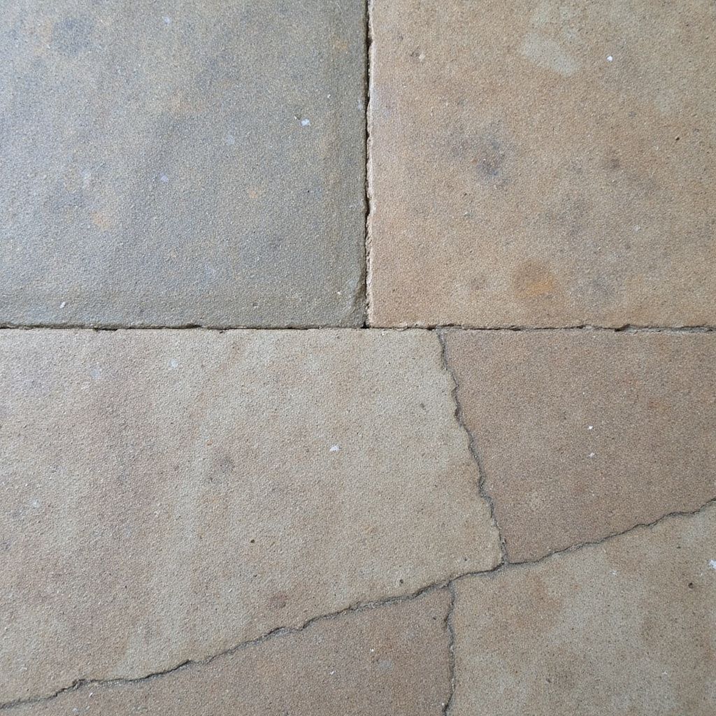 Construction worker inspecting a sidewalk crack in front of a building; wearing a yellow hard hat and orange vest.