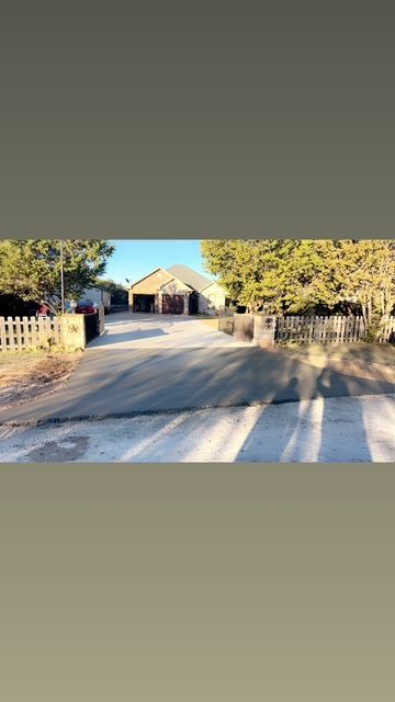 Long driveway leads to a light-colored building with a dark roof. Trees and a fence are on either side.
